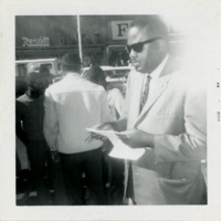 Man in sunglasses holds paper with people in the background, 1964